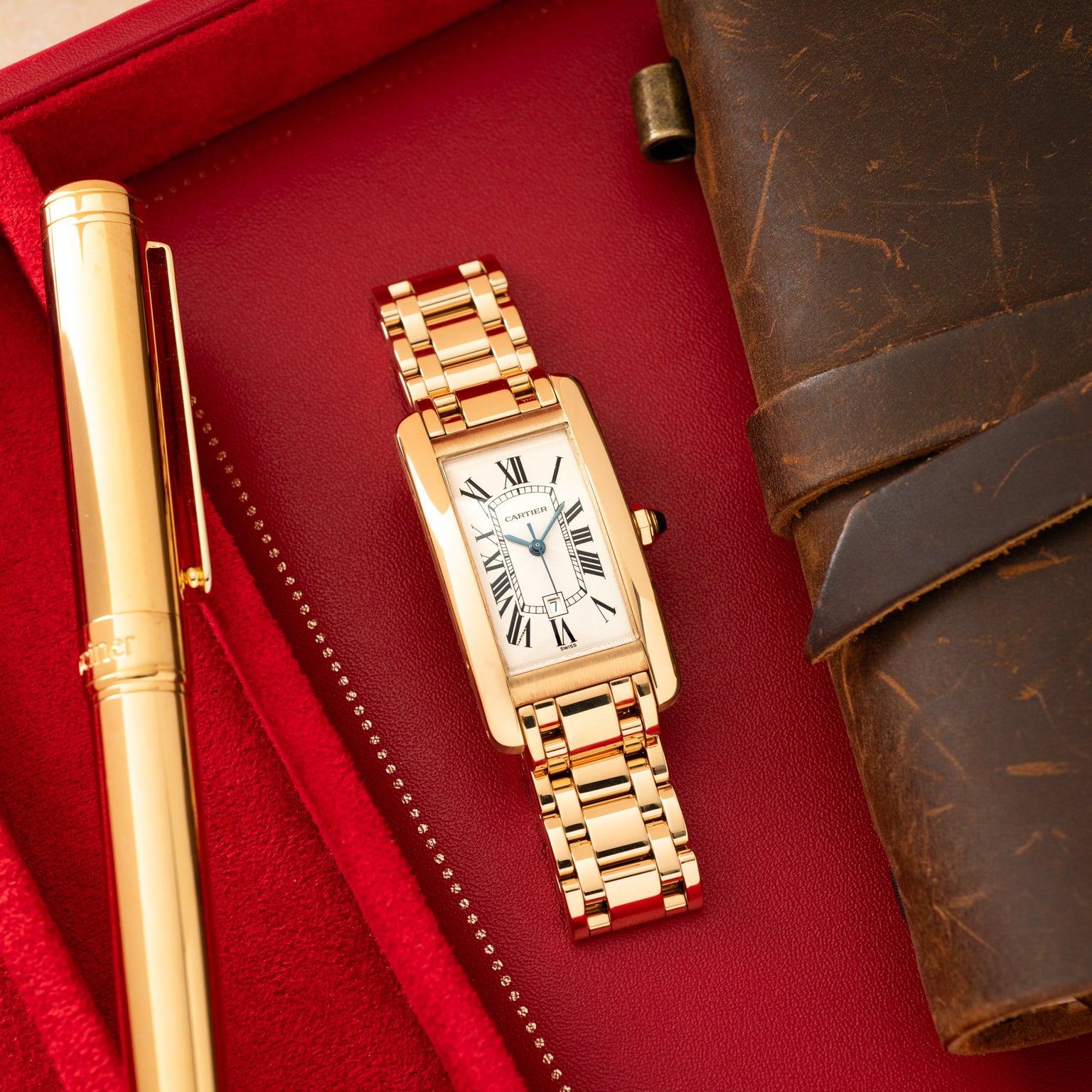 Gold-tone rectangular watch with white dial and Roman numerals on red fabric next to a pen and leather accessories.