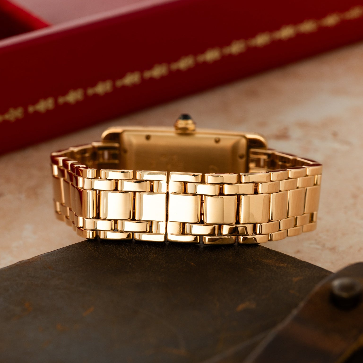 Gold-tone rectangular watch with linked bracelet and crown detail, displayed on dark stone with red box background.