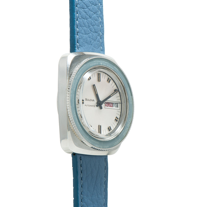 Bulova automatic watch with square stainless steel case, white dial, day-date window, and blue leather strap.
