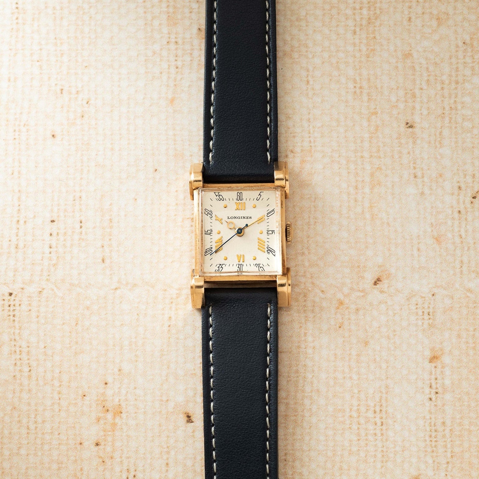 Gold-tone rectangular watch with cream dial, Roman numerals, and black leather strap with white stitching.
