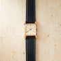 Gold-tone rectangular watch with cream dial, Roman numerals, and black leather strap with white stitching.