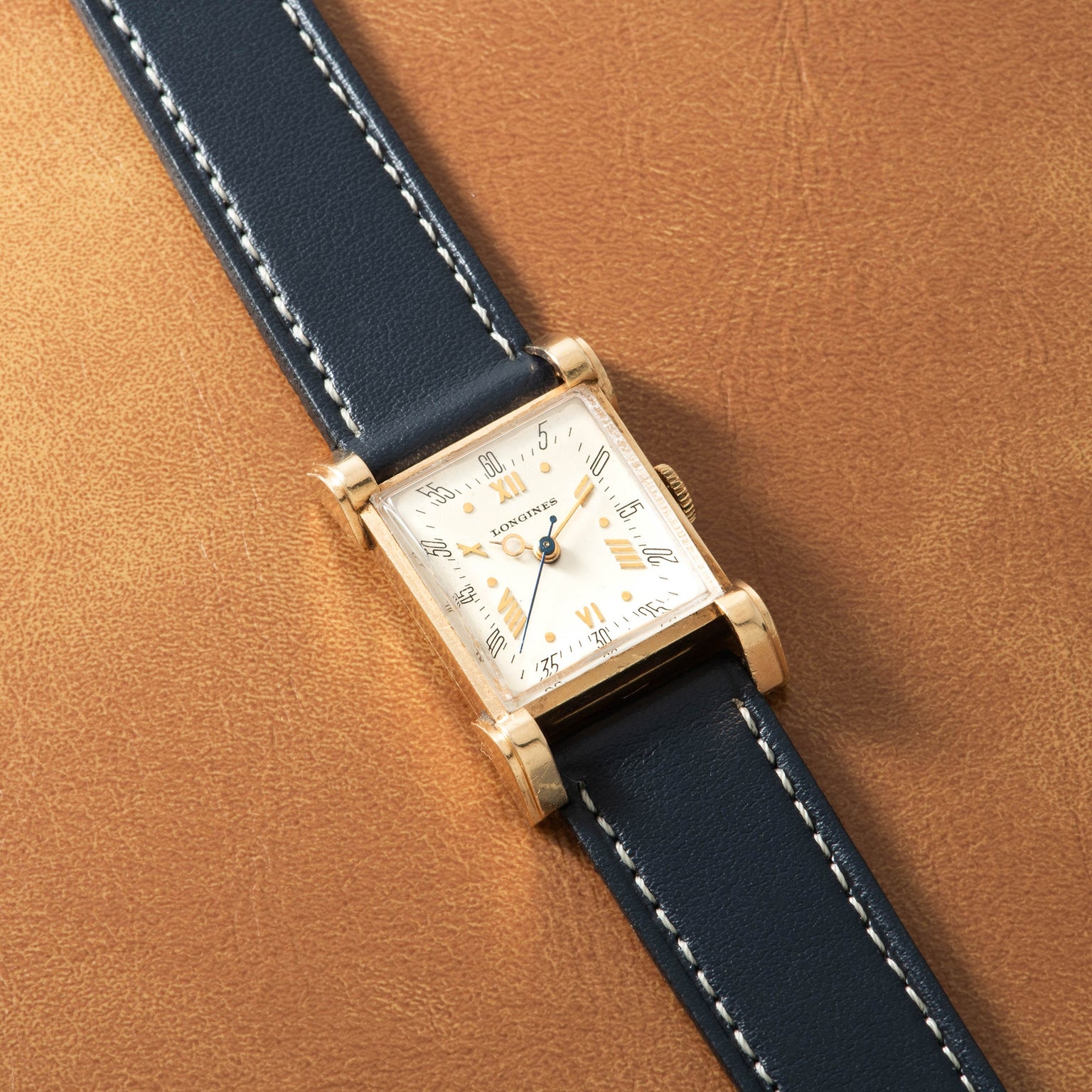Longines Tank watch with square gold case, white dial, hooded lugs, and dark blue leather strap with white stitching.