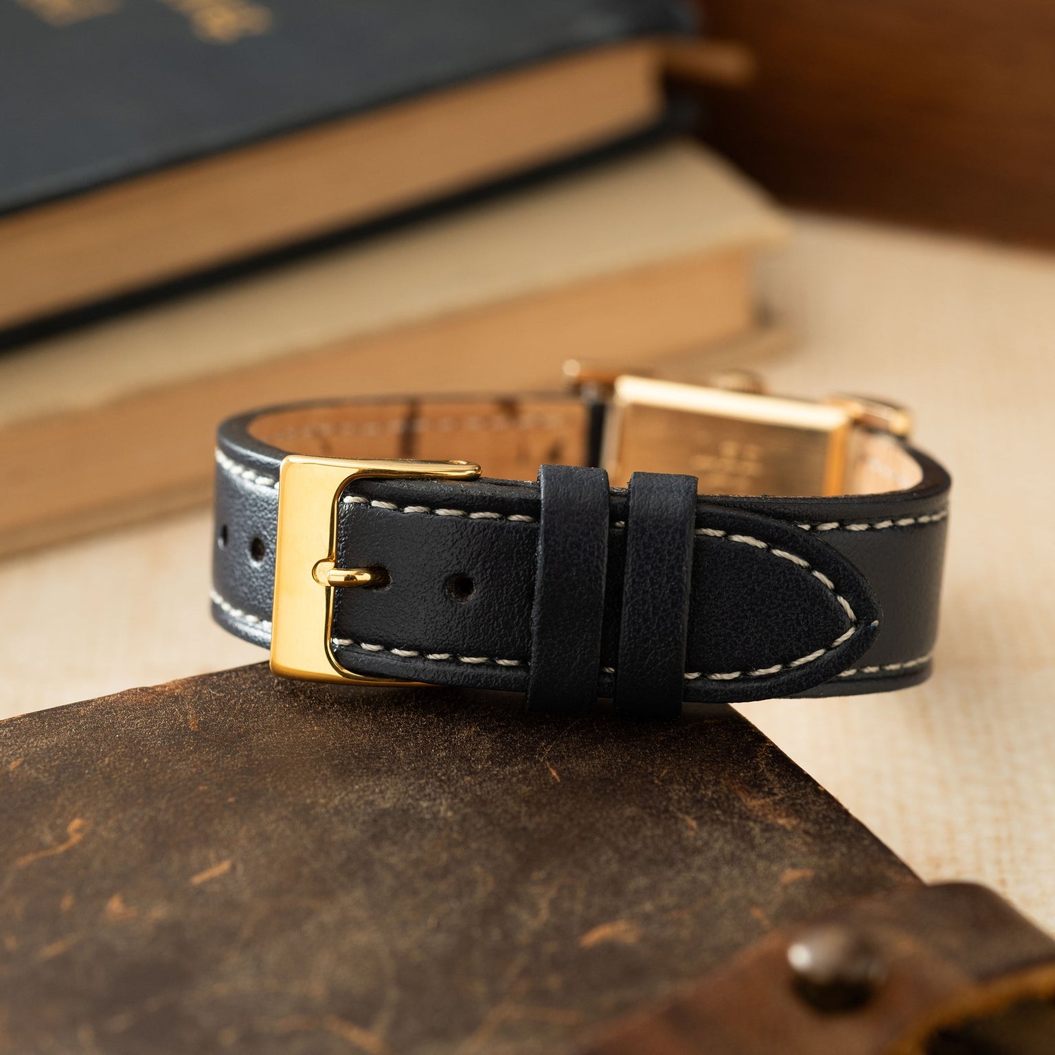 Gold-toned rectangular watch case with black leather strap, white stitching, and gold buckle on tan surface.
