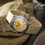 Vintage military watch with cream dial, Arabic numerals, and tan fabric strap resting on khaki fabric.