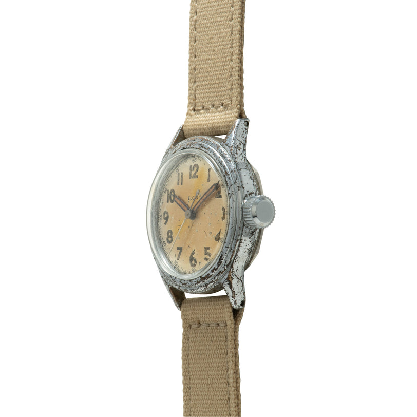 Vintage Elgin wristwatch with cream dial, Arabic numerals, weathered metal case, and tan fabric strap.