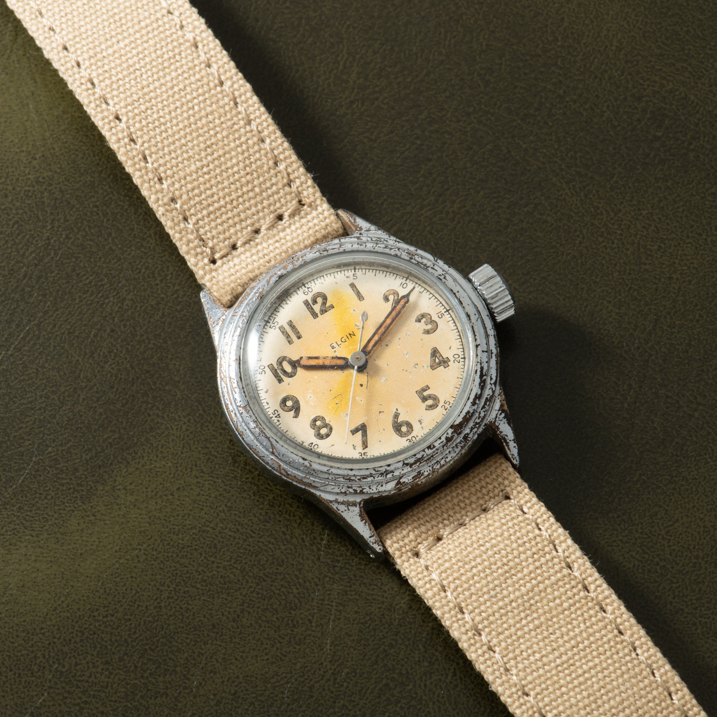 Elgin watch with cream dial, Arabic numerals, and worn steel case on beige canvas strap against olive background.