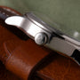 Stainless steel watch crown with cross emblem on brown leather strap against green background.