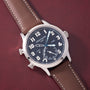 Silver-cased watch with black dial, white numerals, dual time zones marked "HOME" and "LOCAL", and brown leather strap with w
