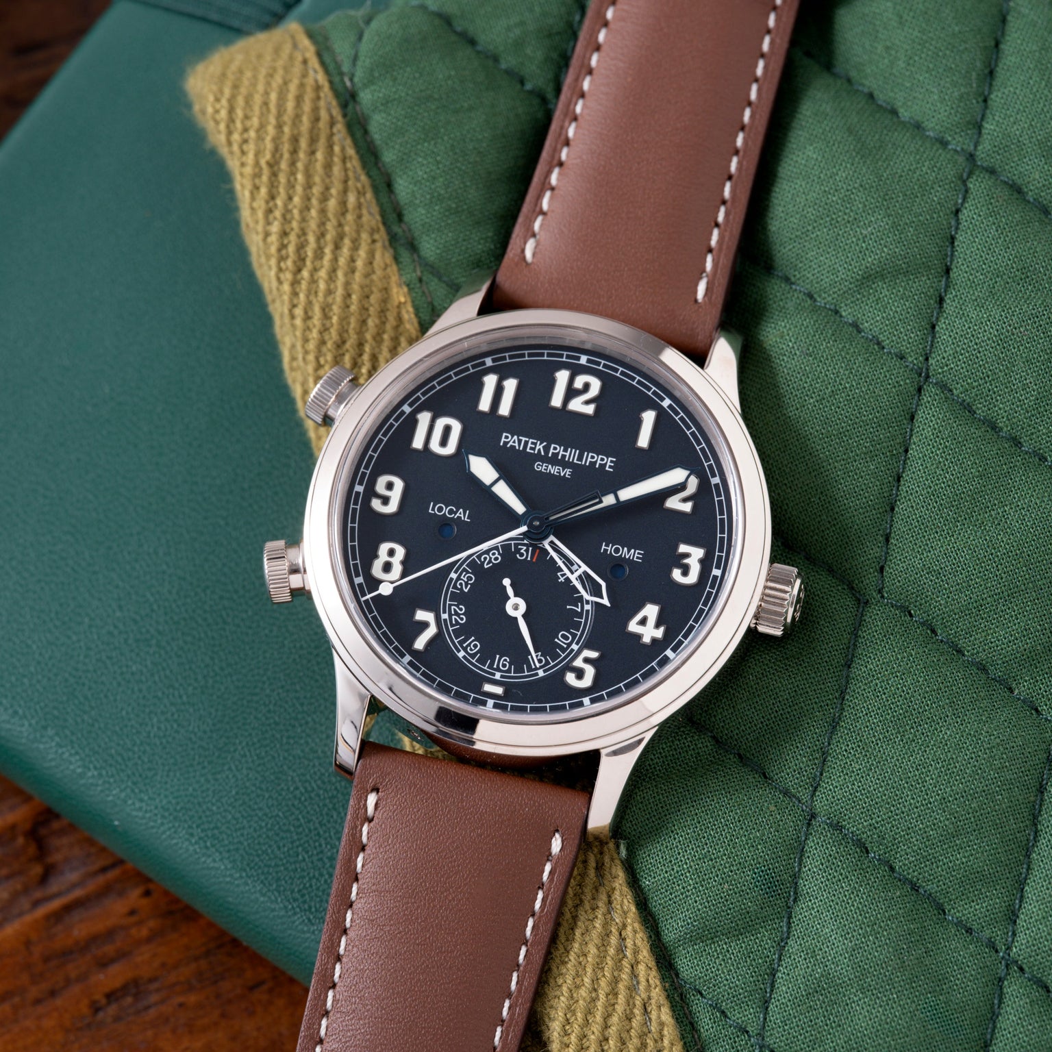 Patek Philippe watch with black dial, dual time zones marked "LOCAL" and "HOME," brown leather strap, displayed on green and