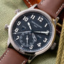 Patek Philippe watch with black dial, Arabic numerals, dual time zone subdial, and brown leather strap on fabric background.
