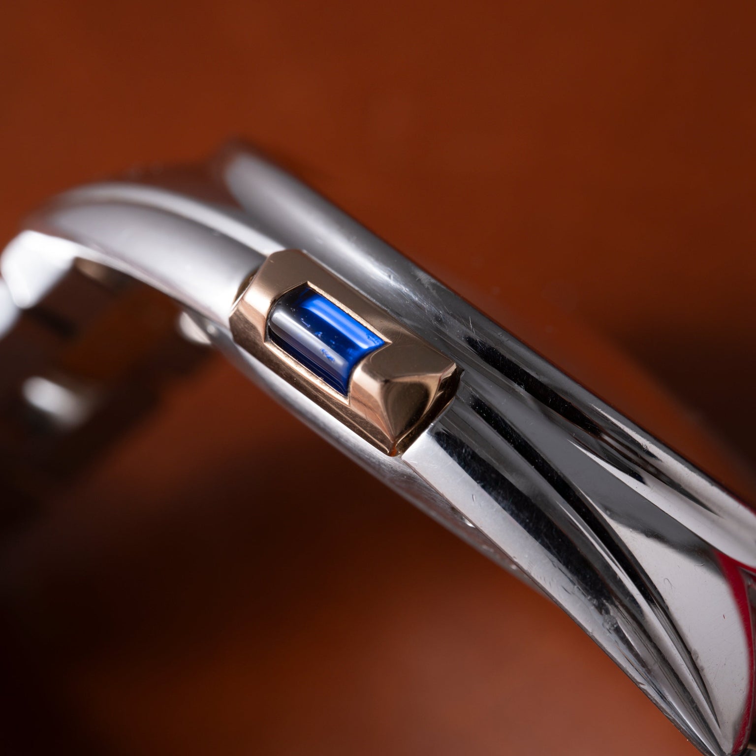 Cartier Clé de Cartier watch with white dial, metal bracelet, and blue sapphire stone on crown against brown background.