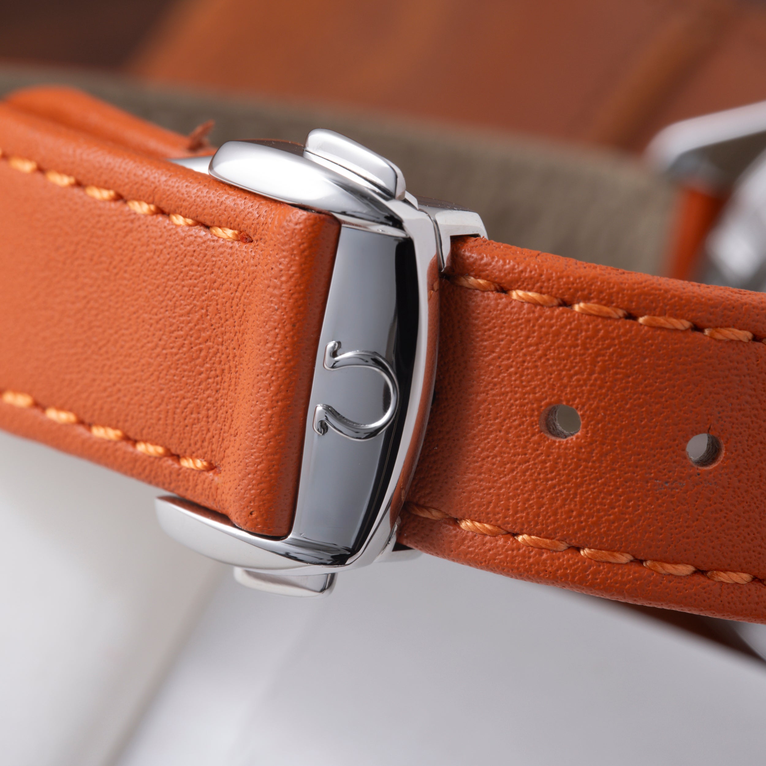 Polished steel watch case with crown detail on orange leather strap with tan stitching.