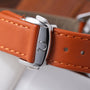 Polished steel watch case with crown detail on orange leather strap with tan stitching.