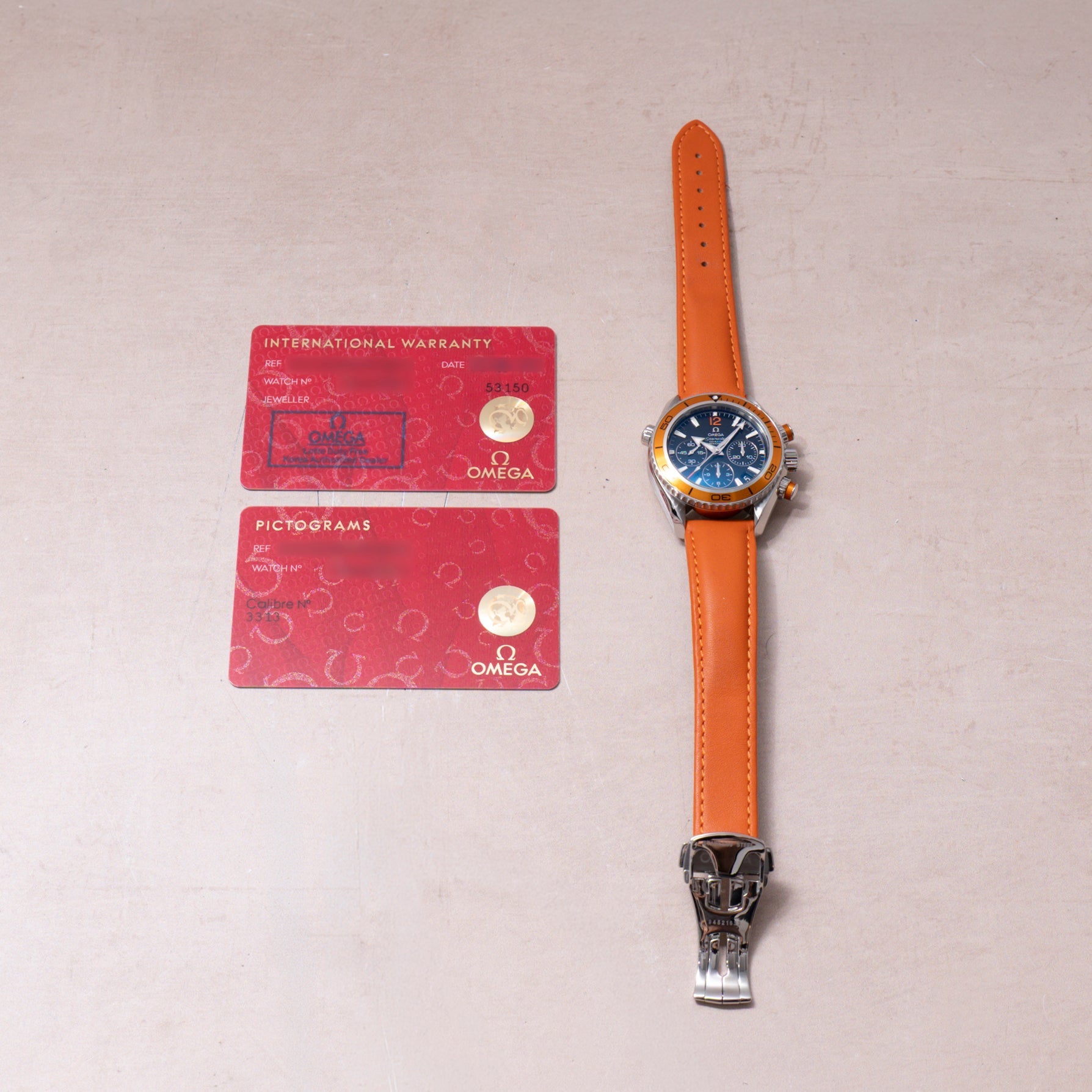 Omega watch with blue dial and orange leather strap, displayed with two red warranty and pictogram cards.