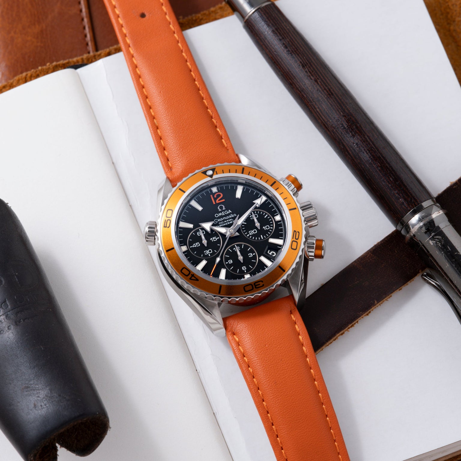 Stainless steel chronograph watch with orange bezel and orange leather strap on white background with leather accessories.