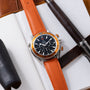 Stainless steel chronograph watch with orange bezel and orange leather strap on white background with leather accessories.
