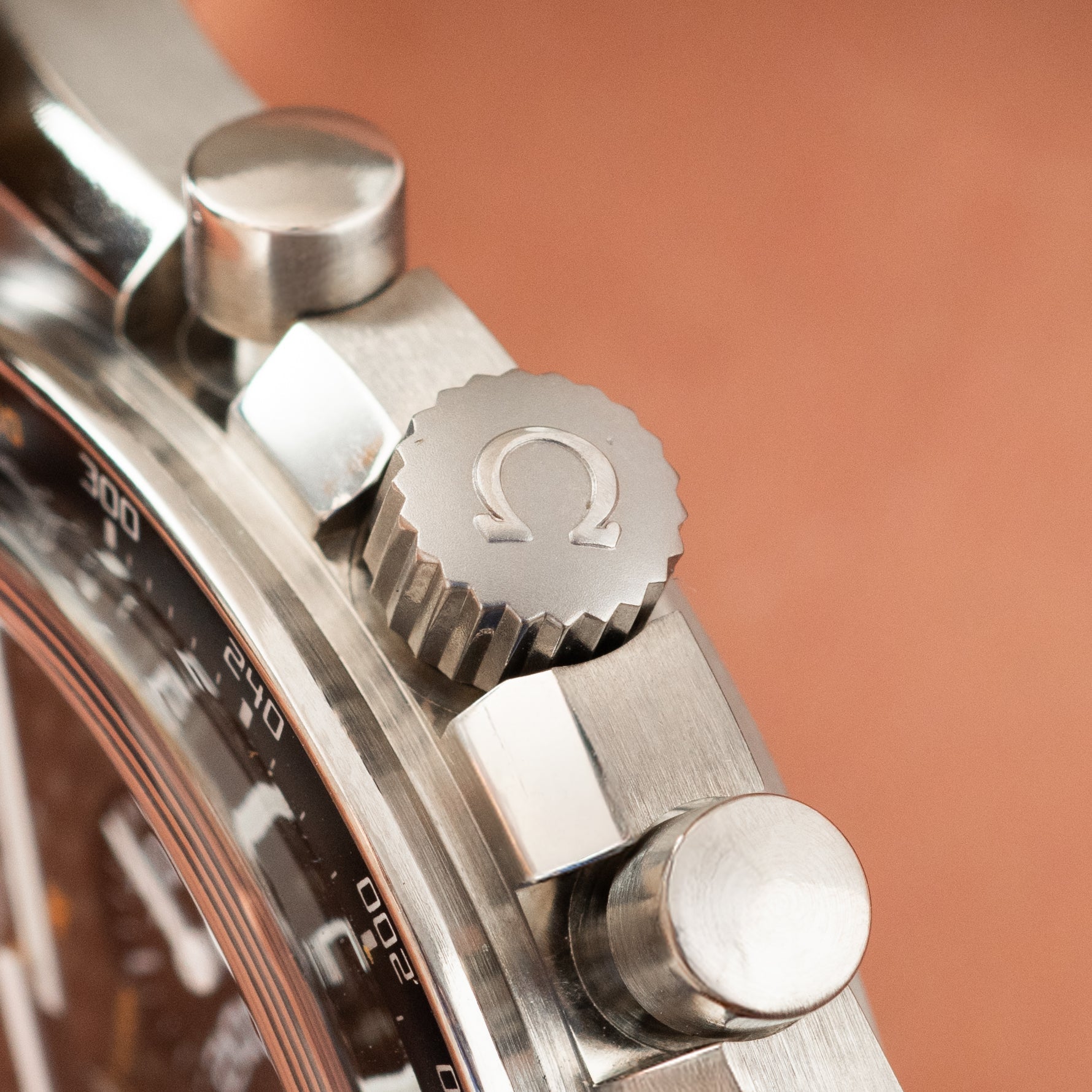Titanium watch crown and pushers with Omega symbol engraved on crown, shown against coral background.