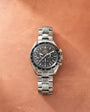 Silver-tone Omega Speedmaster chronograph watch with black dial, tachymeter bezel, and metal bracelet on tan background.