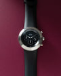 Black chronograph watch with silver round case, black dial with subdials, and black rubber strap.
