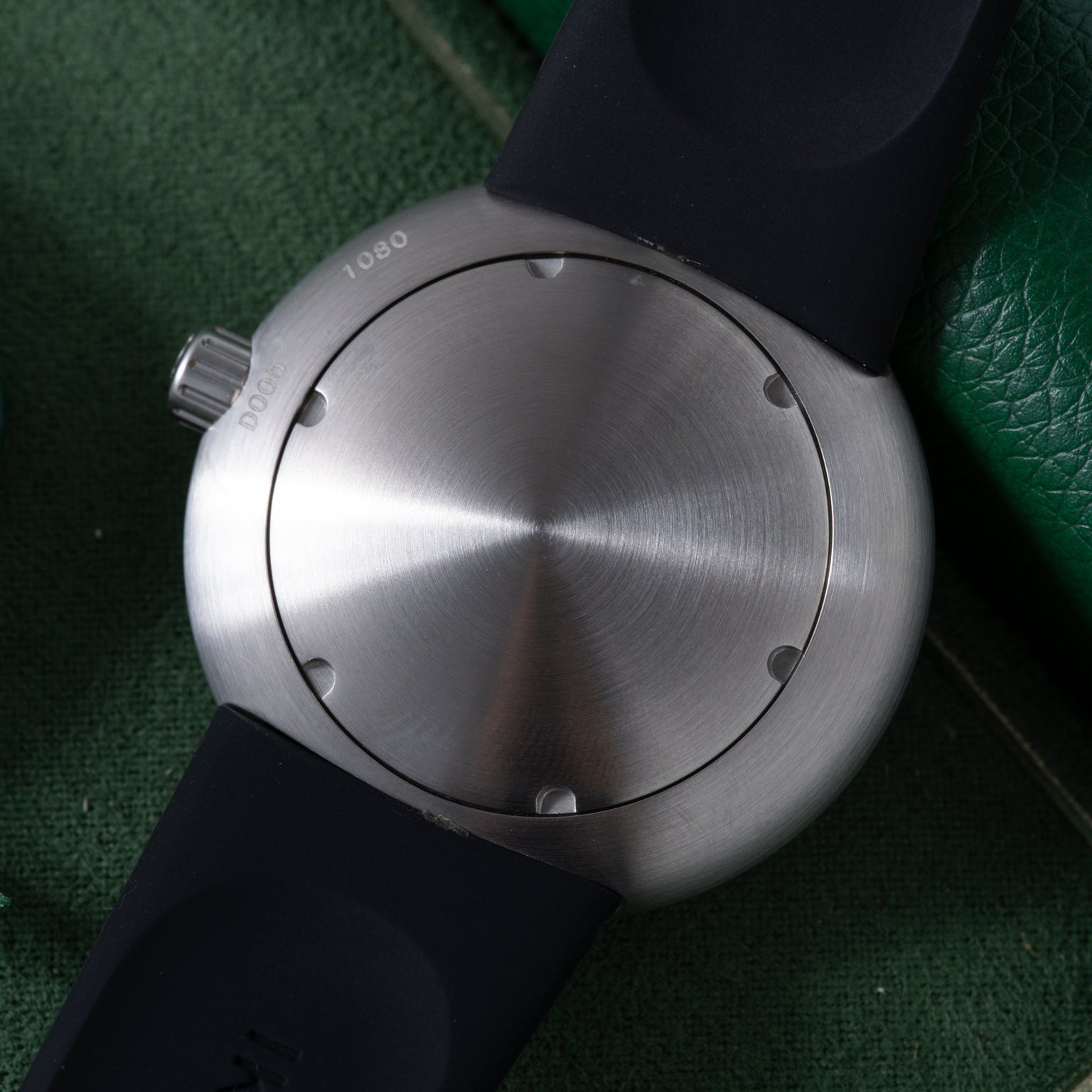 Brushed stainless steel watch face with black rubber strap and crown, displayed on green textured background.