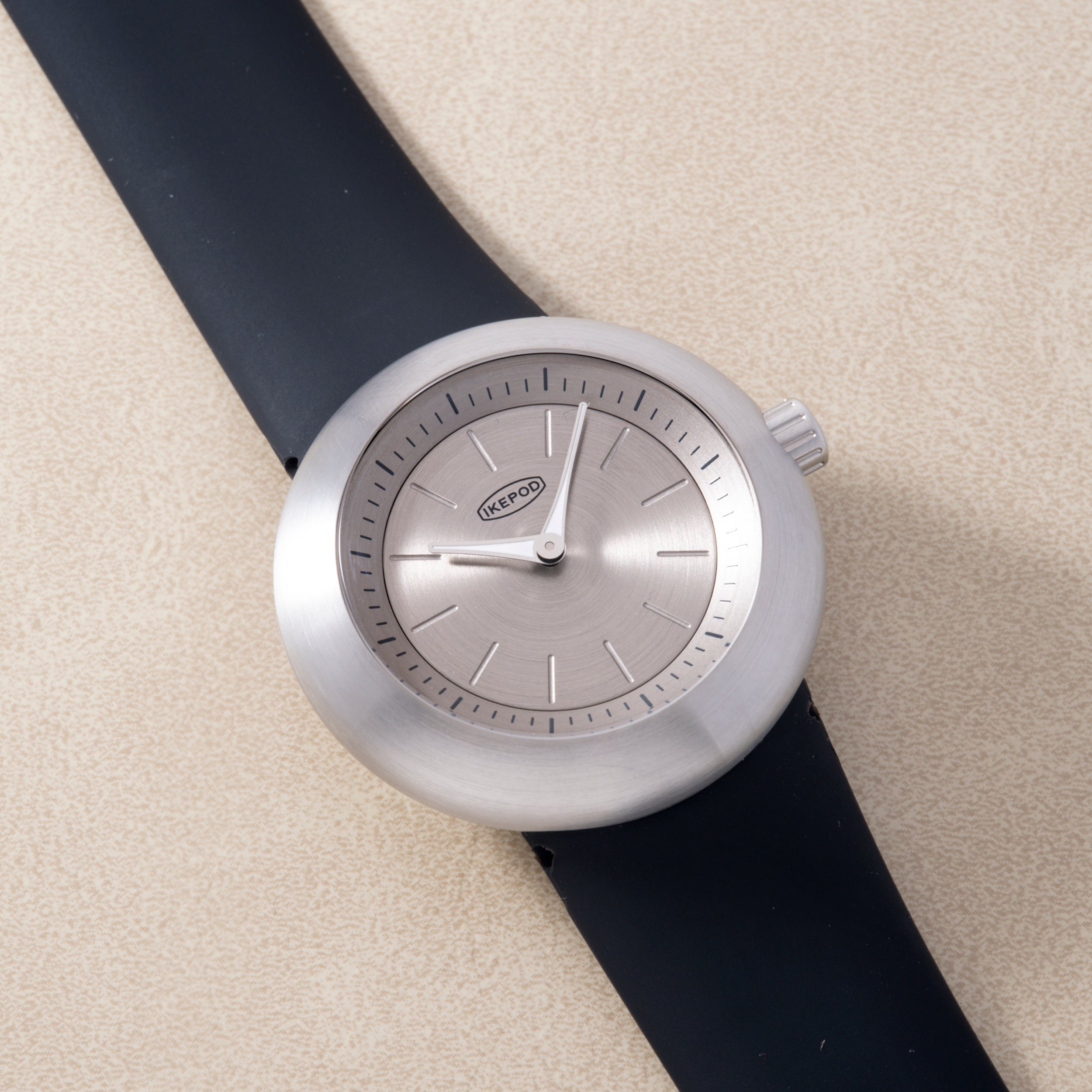 Round silver watch with gray dial, hour markers, and black rubber strap against beige background.