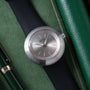 Ikepod Duopod watch with round brushed steel case, grey dial, and dark navy leather strap on green textile background.