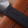 Black Ikepod strap with gold dots on tan leather band next to silver watch face.