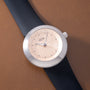 White round watch with textured gold dial, black leather strap, and hour markers on beige background.