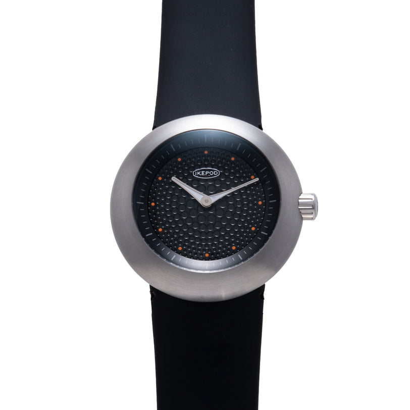 Ikepod watch with brushed metal case, black dial featuring orange dot markers, white hands, and black rubber strap.