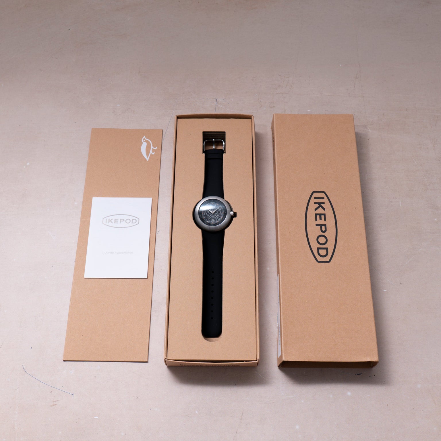 Ikepod Duopod Dots 004 watch with black leather strap and silver case displayed in open kraft packaging.