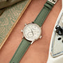 Silver-tone watch with cream dial, Arabic numerals, and green fabric strap on arm with map and book nearby.