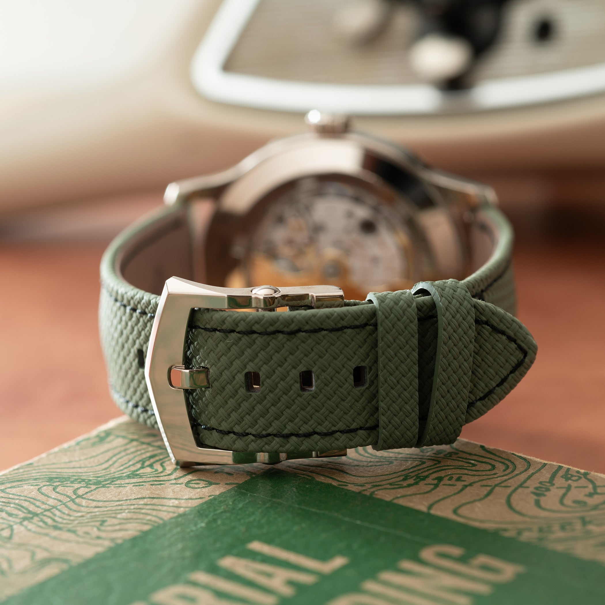 Stainless steel watch with gray dial and olive green woven fabric strap, displayed on branded packaging.