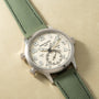 Silver-tone watch with cream dial, Arabic numerals, dual time zones marked "HOME" and "LOCAL," and green fabric strap.