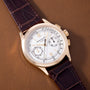 Gold-tone chronograph watch with white dial, Roman numerals, subdials, and dark purple crocodile leather strap.