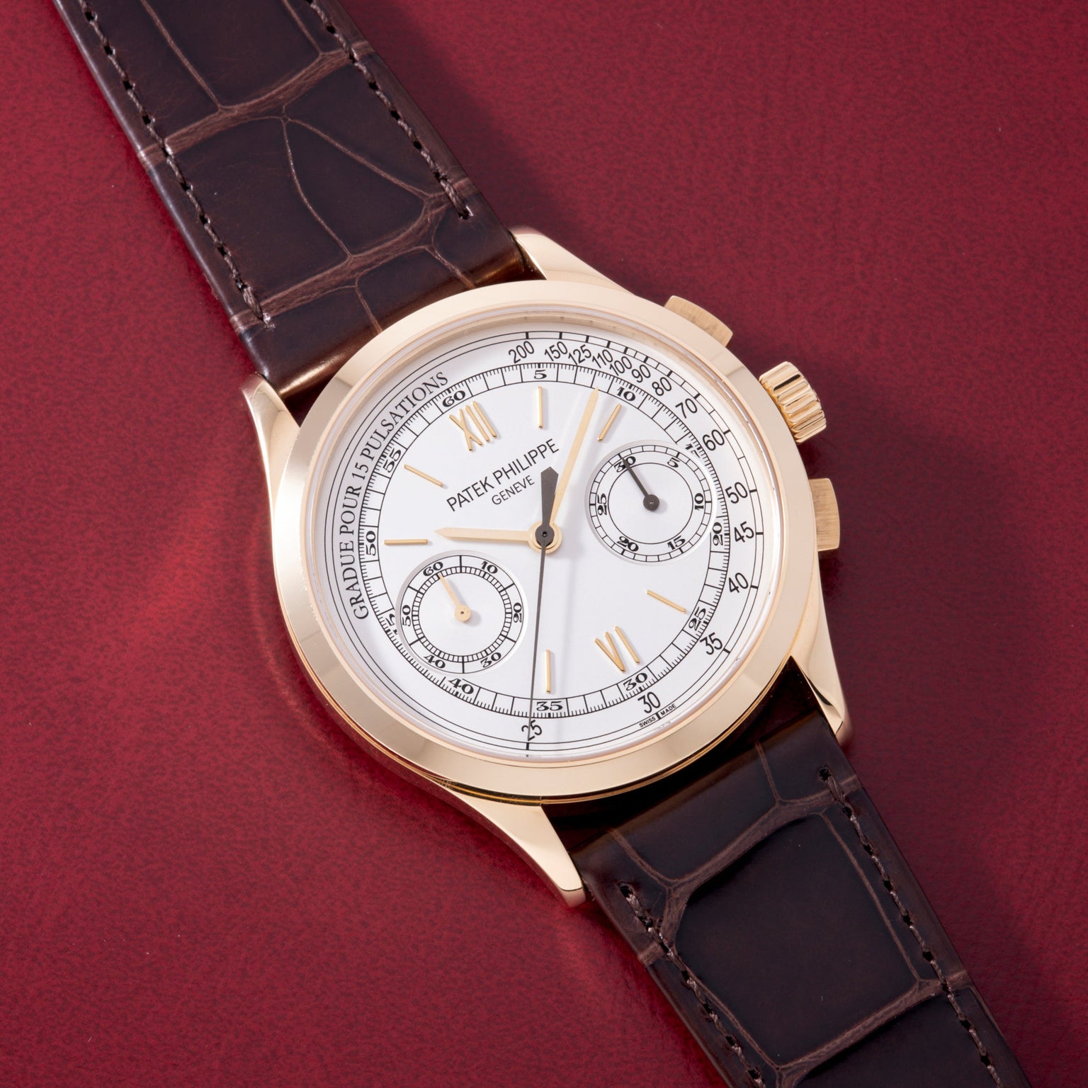 Gold-tone chronograph watch with white dial, two subdials, Roman numerals, and dark crocodile leather strap on burgundy backg