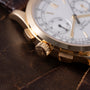 Gold chronograph watch with white dial, subsidiary dials, and textured crown on dark leather surface.
