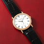 Gold-tone watch with white dial, Roman numerals, subdial, and black crocodile leather strap on red background.