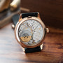 Rose gold watch with skeleton dial displaying visible movement, black leather strap, and regulator-style hour markers.