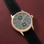 Rose gold watch with gray dial, two subdials, and black leather strap on burgundy background.