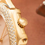 Gold watch with diamond-set bezel, crown, and lugs; pearl dial visible against tan background.