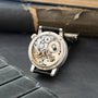 Silver watch with black leather strap displaying exposed mechanical movement with gears, jewels, and balance wheel visible th