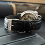 Silver-cased watch with exposed movement visible through display caseback, black leather strap with A. Lange & Söhne buckle.
