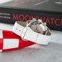 Omega Speedmaster chronograph watch with white leather strap and steel case, displayed with red and white packaging.