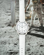 Silver chronograph watch with white dial and white leather strap on lunar surface background.