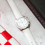 Silver-tone chronograph watch with white dial and white leather strap on concrete surface with red and black branding.