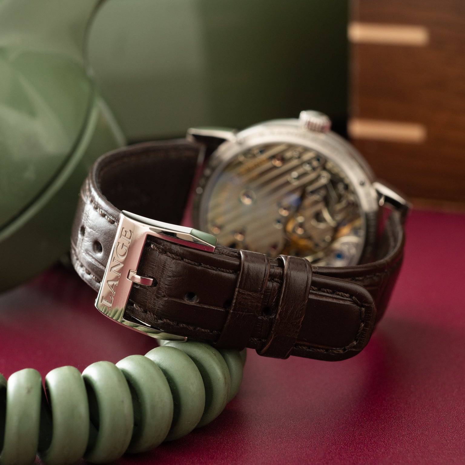A. Lange & Söhne watch with silver case, dark leather strap, and exposed movement dial on burgundy surface.
