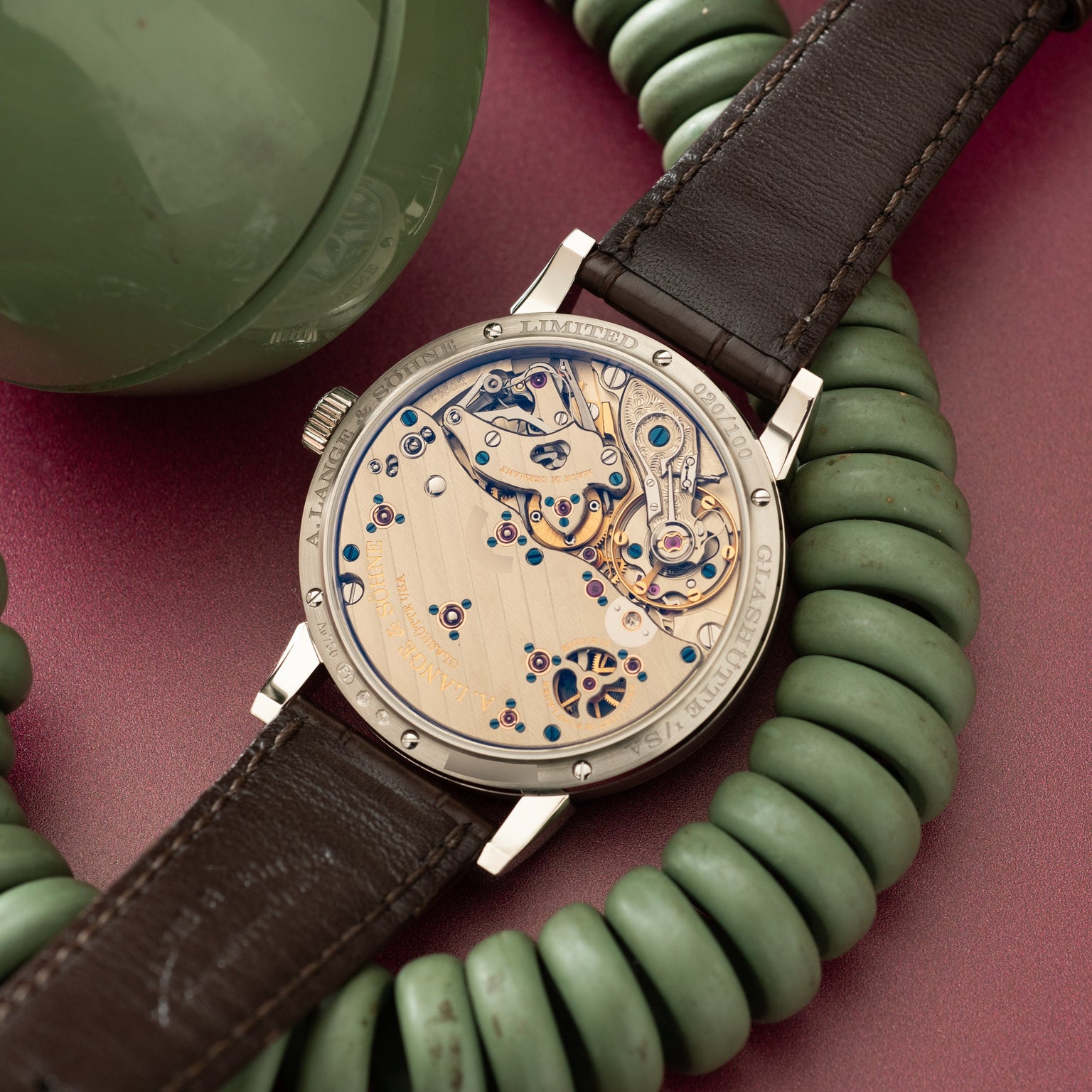 Luxury watch with exposed movement dial, dark brown leather strap, and stainless steel case displayed on burgundy background.