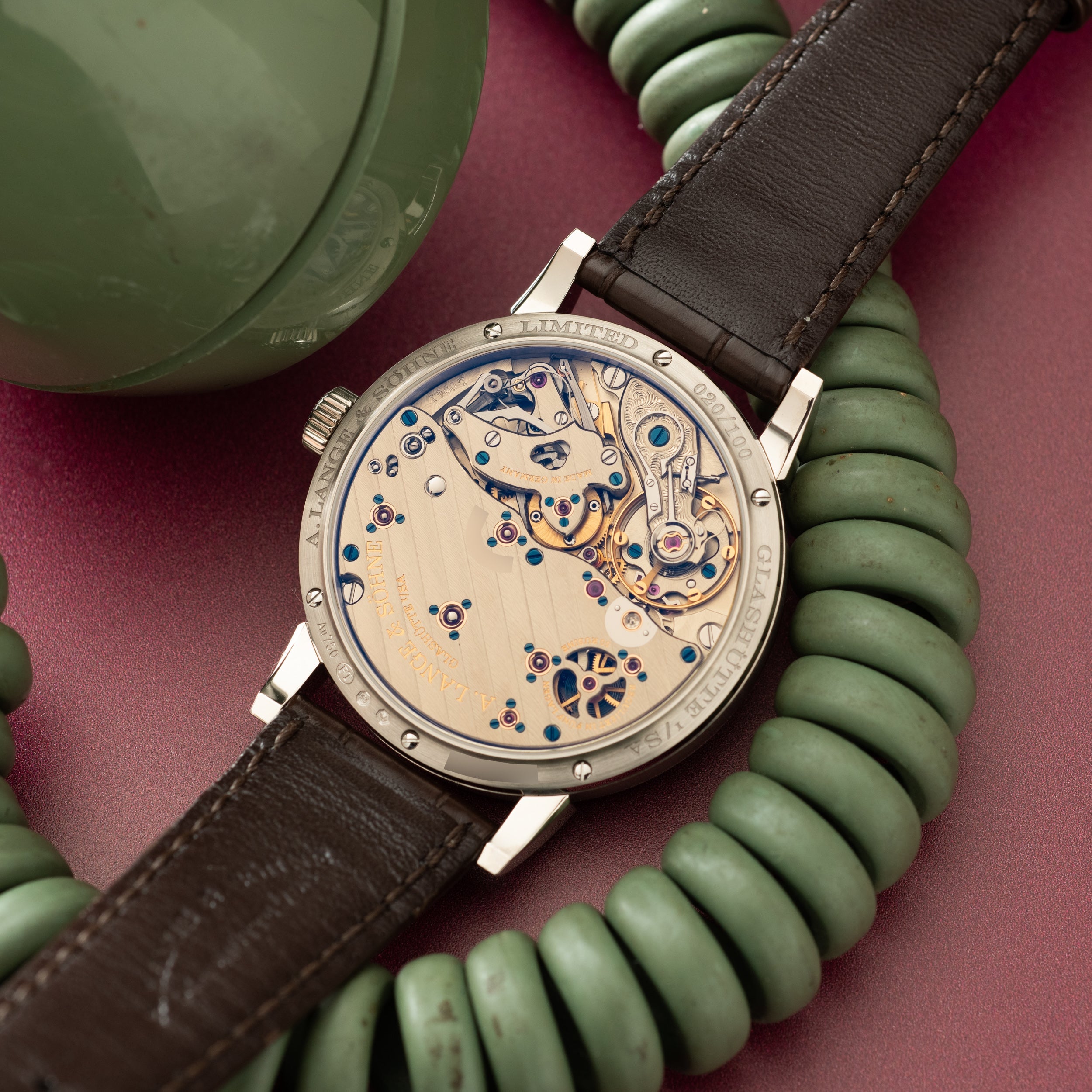 Luxury watch with exposed movement dial, dark brown leather strap, and stainless steel case displayed on burgundy background.