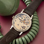 Luxury watch with exposed movement dial, dark brown leather strap, and stainless steel case displayed on burgundy background.