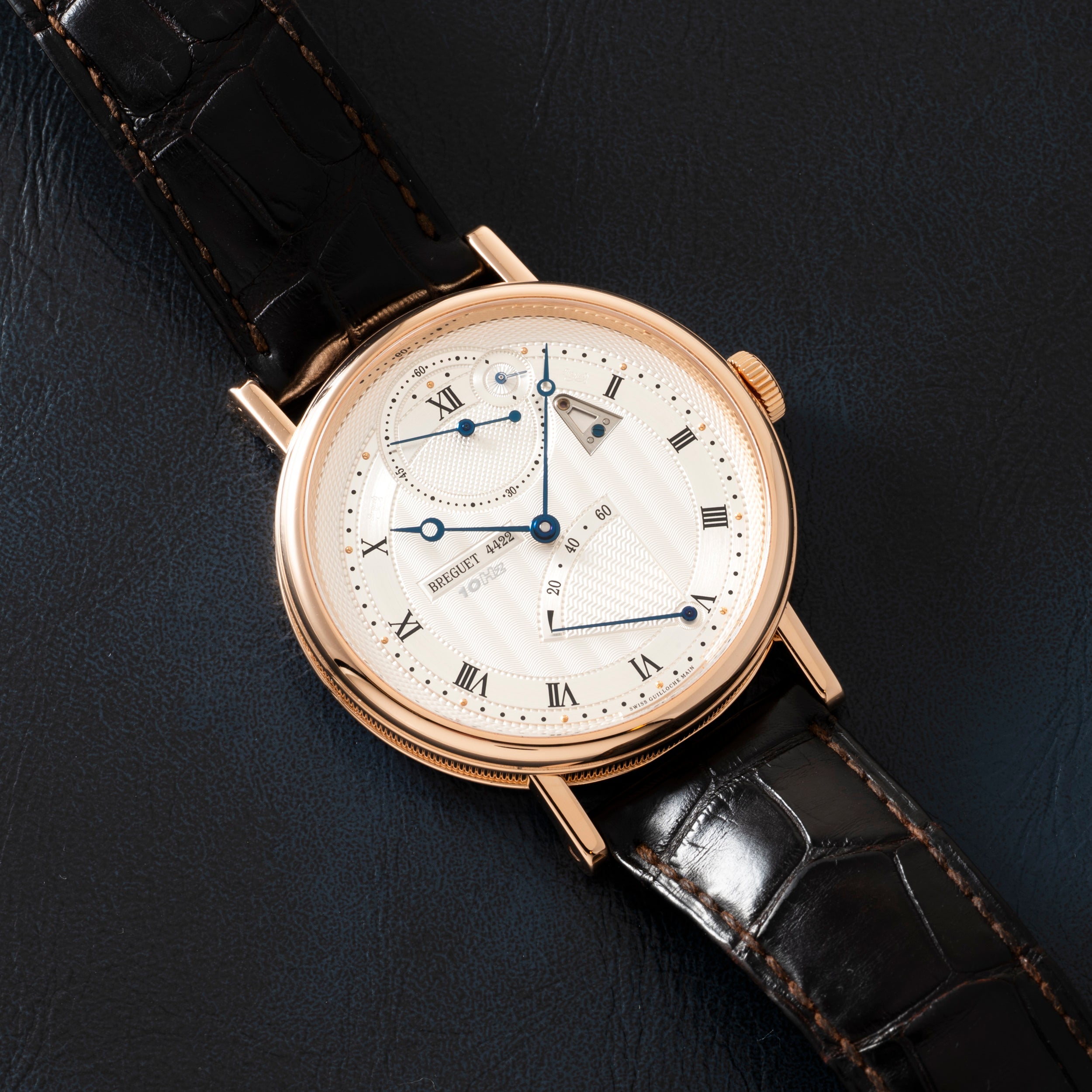 Gold-tone watch with white dial, Roman numerals, multiple subdials, and black leather strap on dark background.